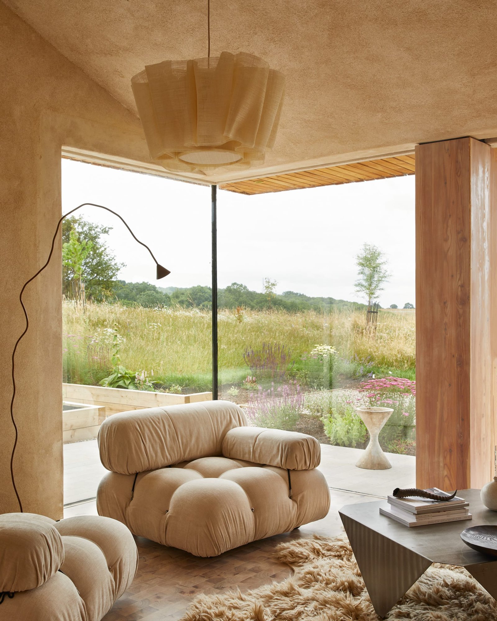 Living room at The Makers Barn with clay-plastered walls and ceiling, a Camaleonda chair and full-height glazing onto a wildflower meadow.
