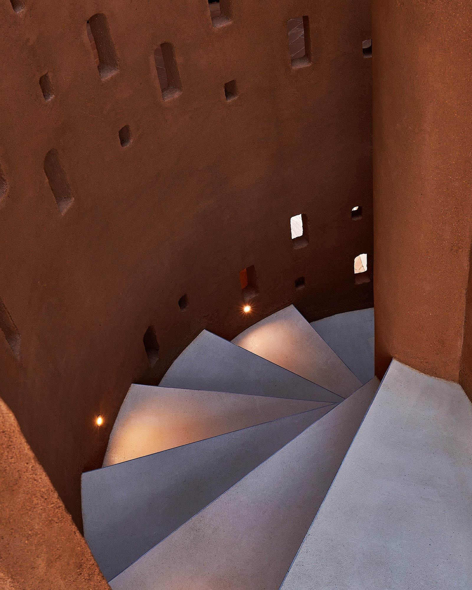 Looking down the spiral staircase at Villa 515: terracotta clay plaster wall pierced with openings above pale grey treads.