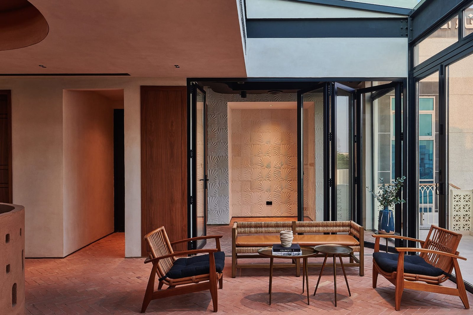 Courtyard lounge at Villa 515 with folding glazing, rattan seating and clay plaster walls meeting a carved tile panel.
