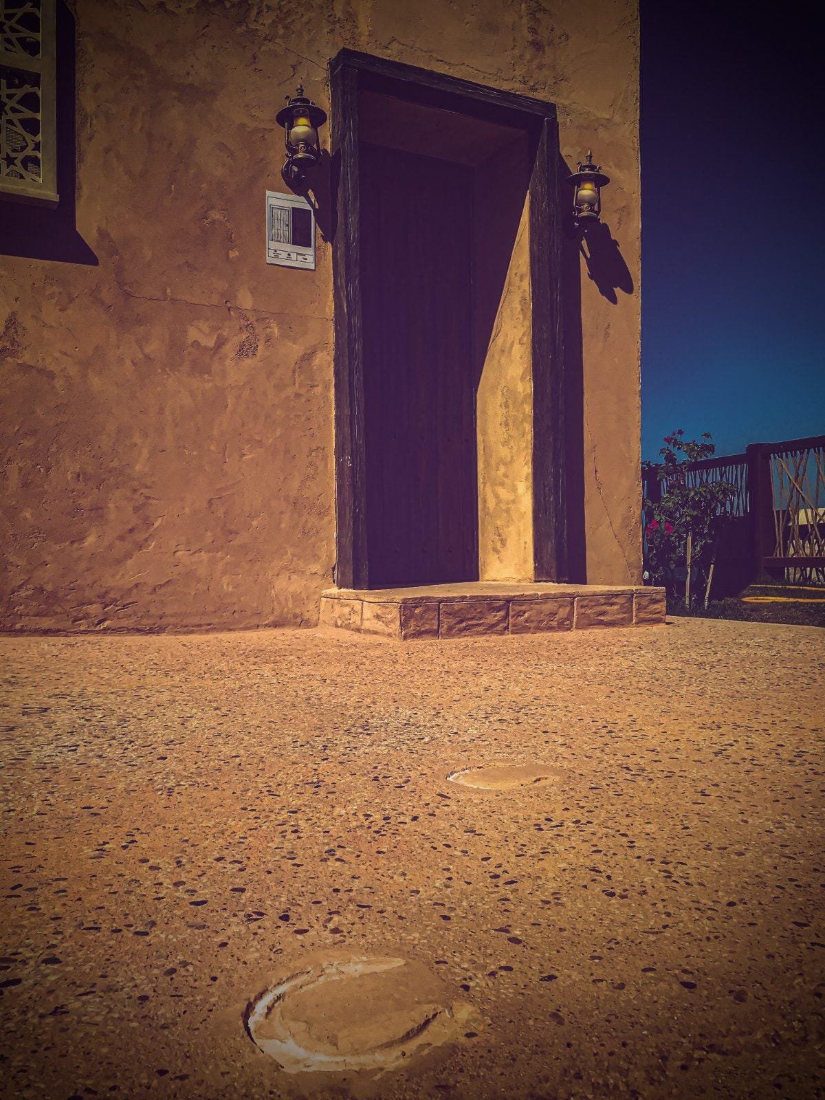 Earthen-plastered heritage building with timber doorway and lantern sconces above an ochre exposed aggregate court with imprinted horseshoe prints
