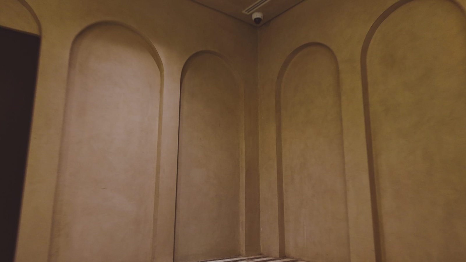 Dim corner of tall arched recessed panels in tan polished plaster, ceiling security camera above, striped tile floor just