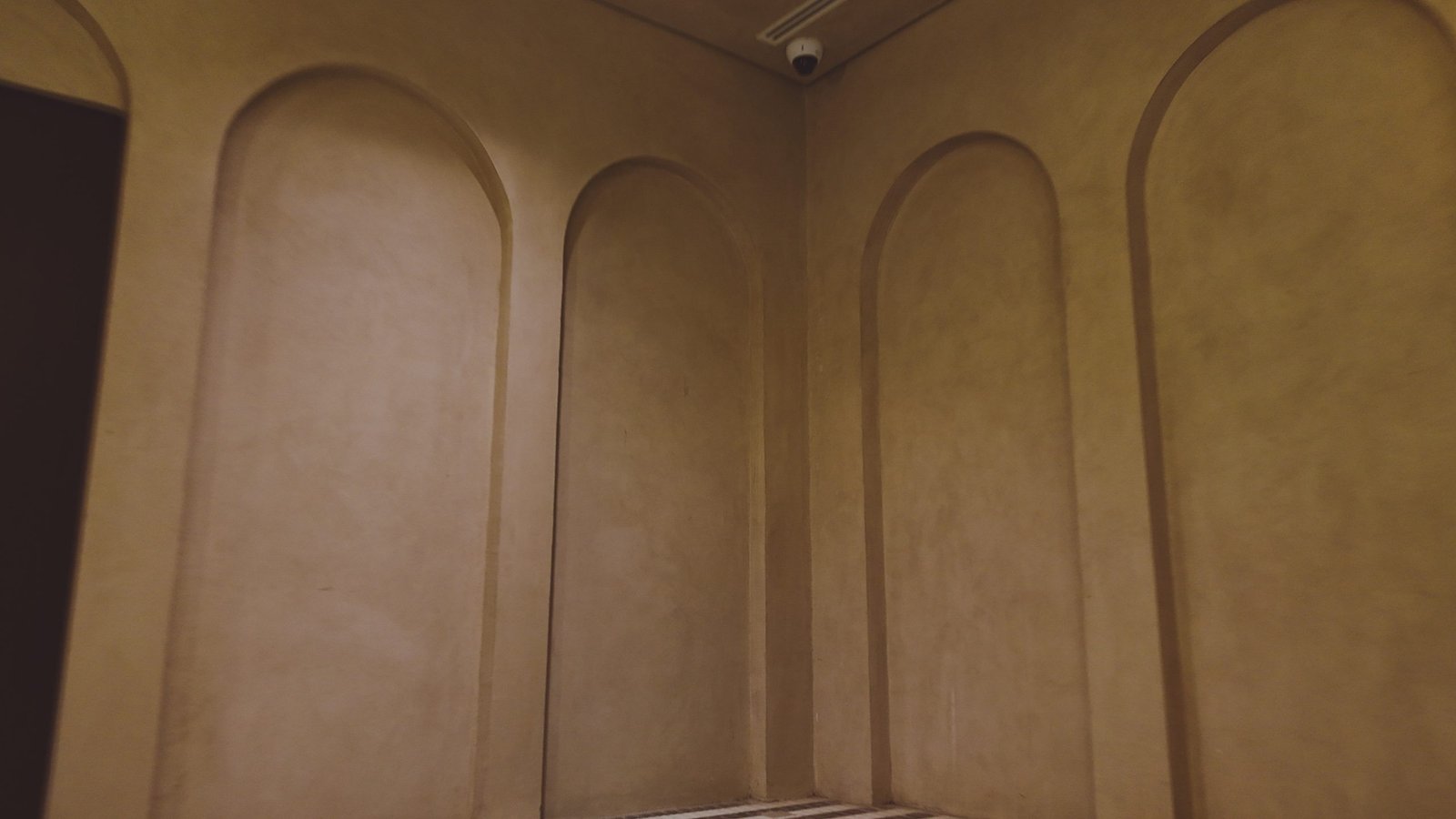 Dim corner of tall arched recessed panels in tan polished plaster, ceiling security camera above, striped tile floor just