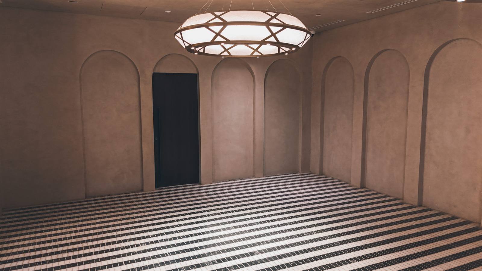 Room lined with blind-arch tadelakt wall panels, black-and-white striped mosaic floor and a geometric ring chandelier over a
