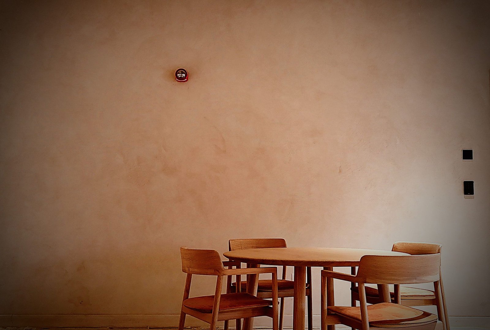 Cafe round oak table with four chairs against an expansive smooth blush-pink plaster wall, red fire-alarm bell mounted high