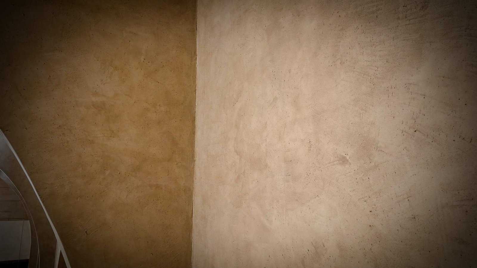 Corner junction of two polished plaster walls, ochre left and pale sand right, white spiral stair rail glimpsed lower left.