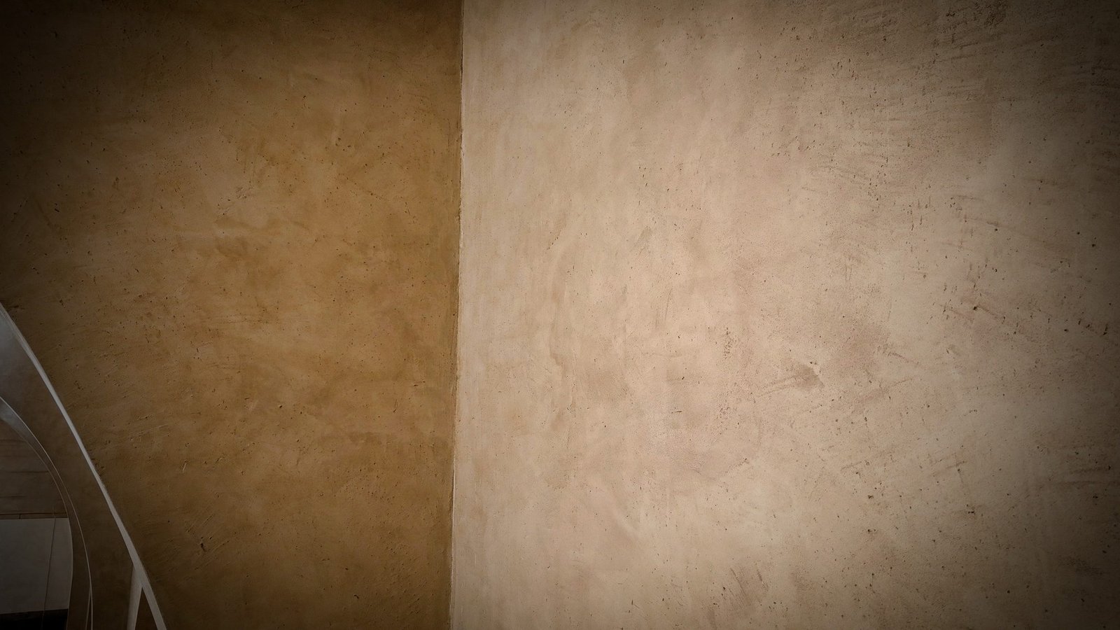 Corner junction of two polished plaster walls, ochre left and pale sand right, white spiral stair rail glimpsed lower left.