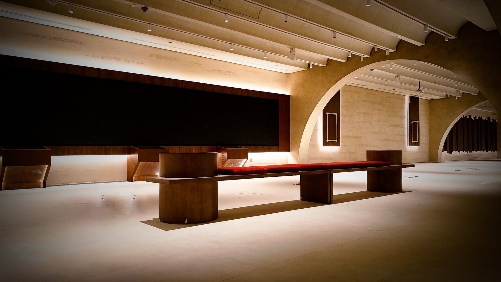 Exhibition hall with a long dark display band, underlit timber benches with red cushions, ribbed cream ceiling with