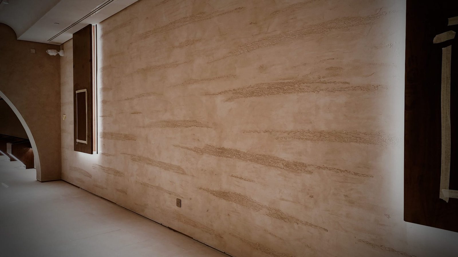 Corridor wall of striated sand Microtadelakt flanked by edge-lit dark timber display panels, arch opening at left, low light.