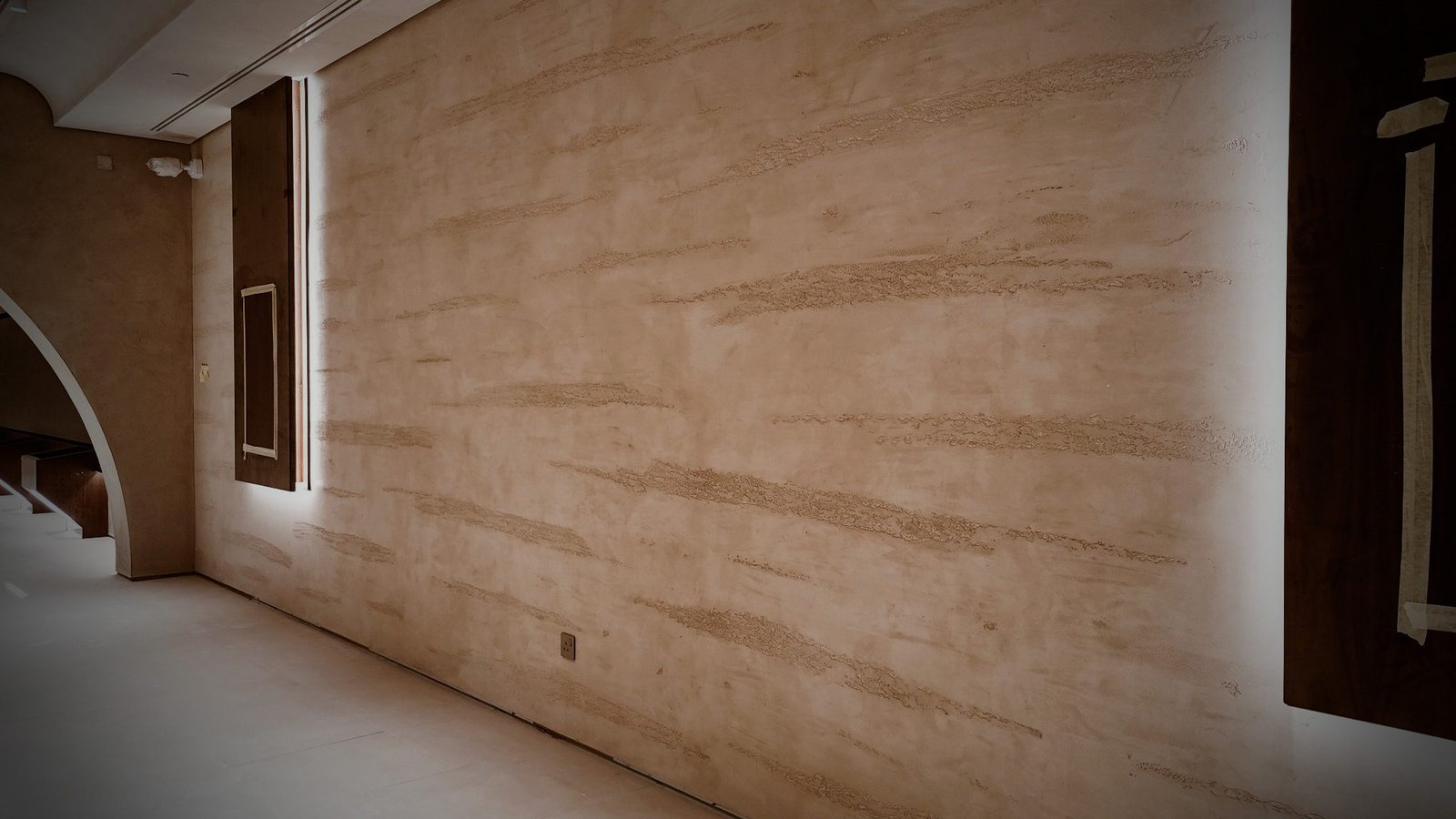 Corridor wall of striated sand Microtadelakt flanked by edge-lit dark timber display panels, arch opening at left, low light.