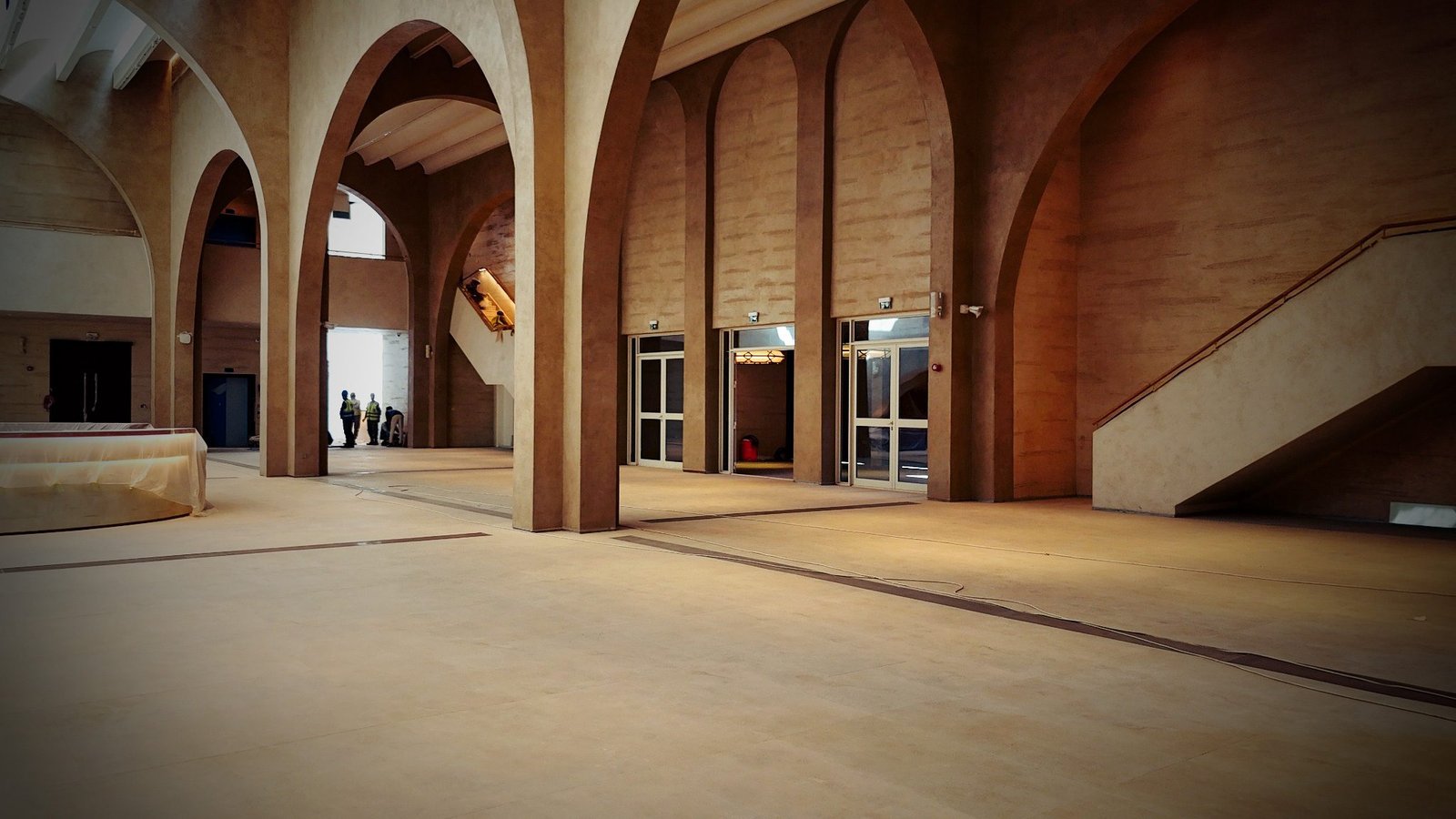 Main hall of arched sand plaster piers and glazed entrance doors, stair at right, distant workers and a sheeted table at