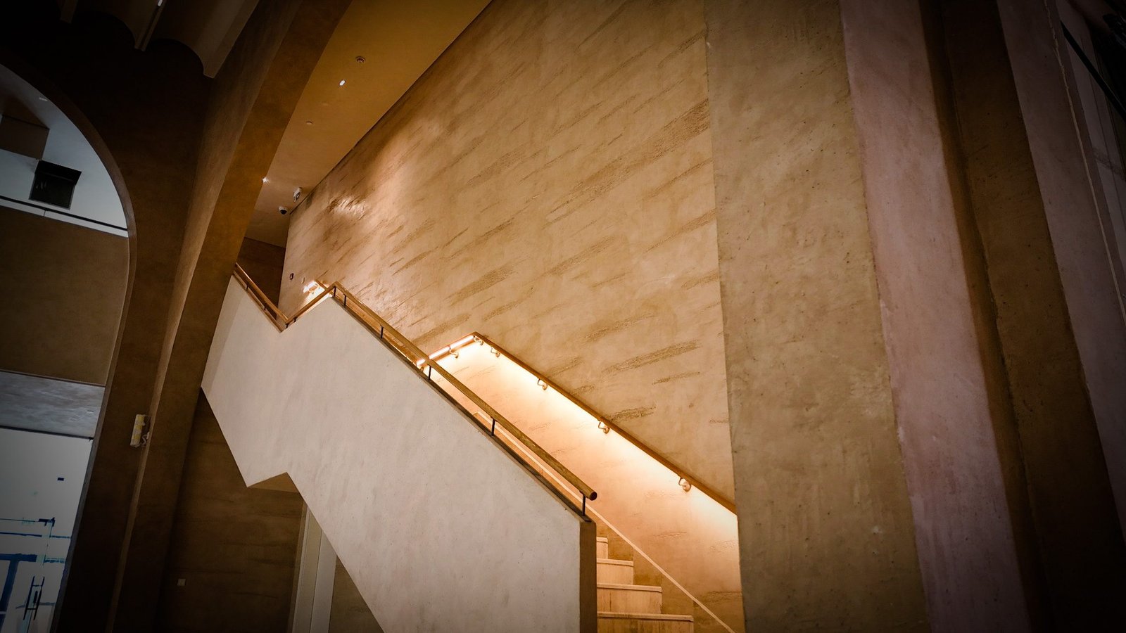 Staircase with pale plaster balustrade and LED-lit timber handrail rising past a tall striated plaster wall, arch glimpsed