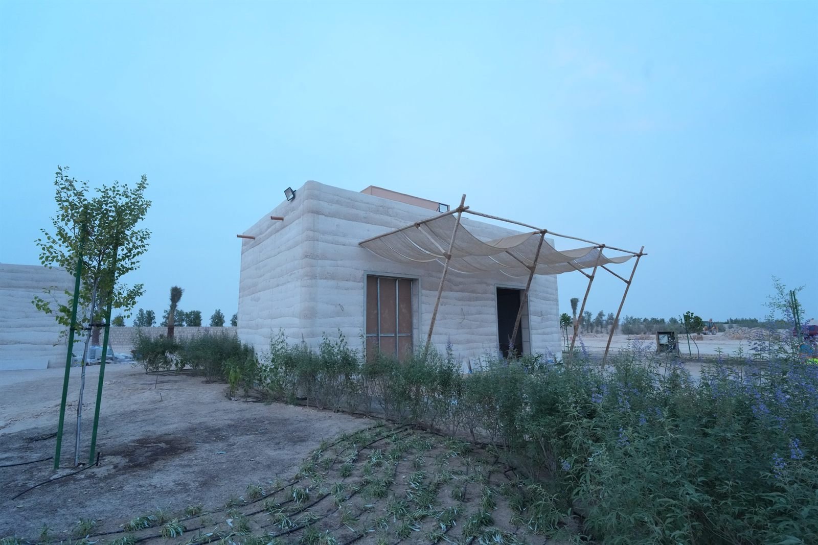 Completed in-situ rammed earth guest building at Al Kaaban Resort, Qatar, layered strata walls at dusk beside a bamboo pergola with draped shade cloth.