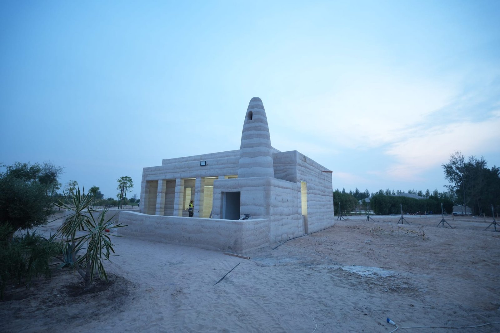 Rammed earth mosque at Al Kaaban Resort, Qatar, its layered conical minaret lit from within at dusk