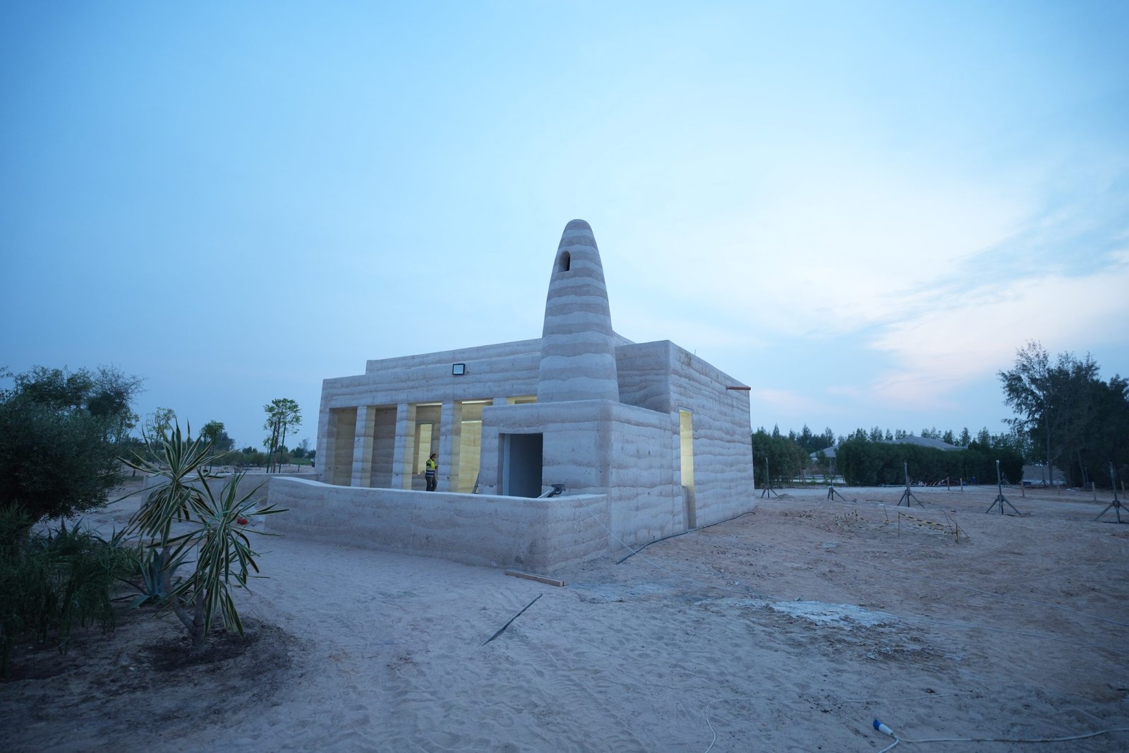 Rammed earth mosque at Al Kaaban Resort, Qatar, its layered conical minaret glowing from warm interior lighting at dusk above a low rammed earth boundary wall.