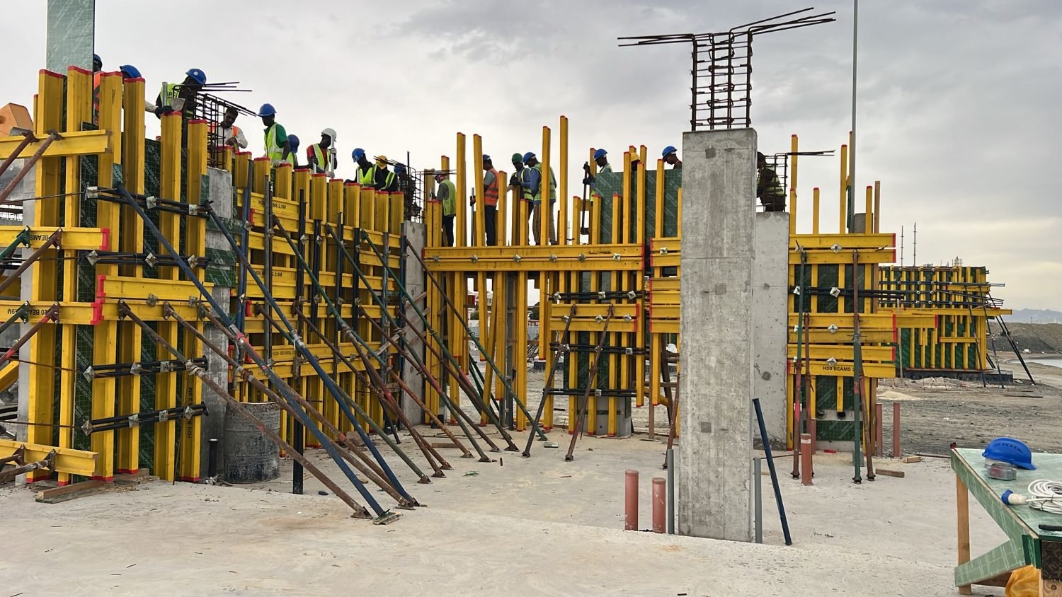 Crews in hi-vis on yellow H20 beam formwork at Kalba Beach, Sharjah, with the Gulf of Oman behind them
