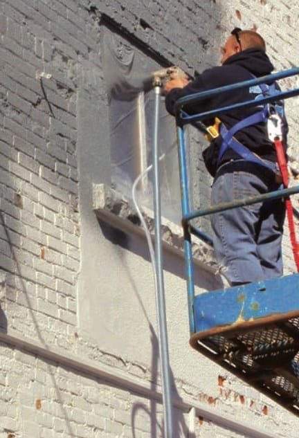 Applicator spraying projected cork onto a painted brick wall from an access platform