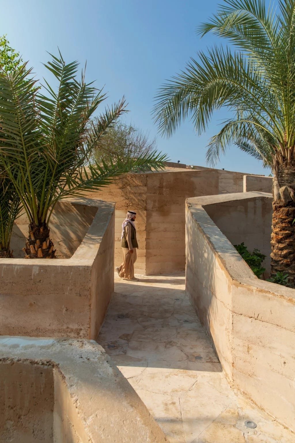 Strata-banded in-situ rammed earth garden walls with date palms, a visitor walking the shaded route between them