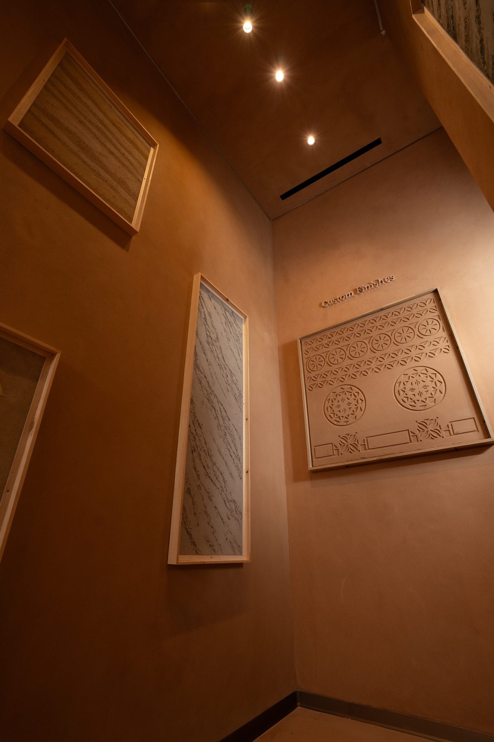 Corner of the Custom Finishes gallery at the Conmarble showroom, framed clay plaster panels on terracotta walls under warm spots.