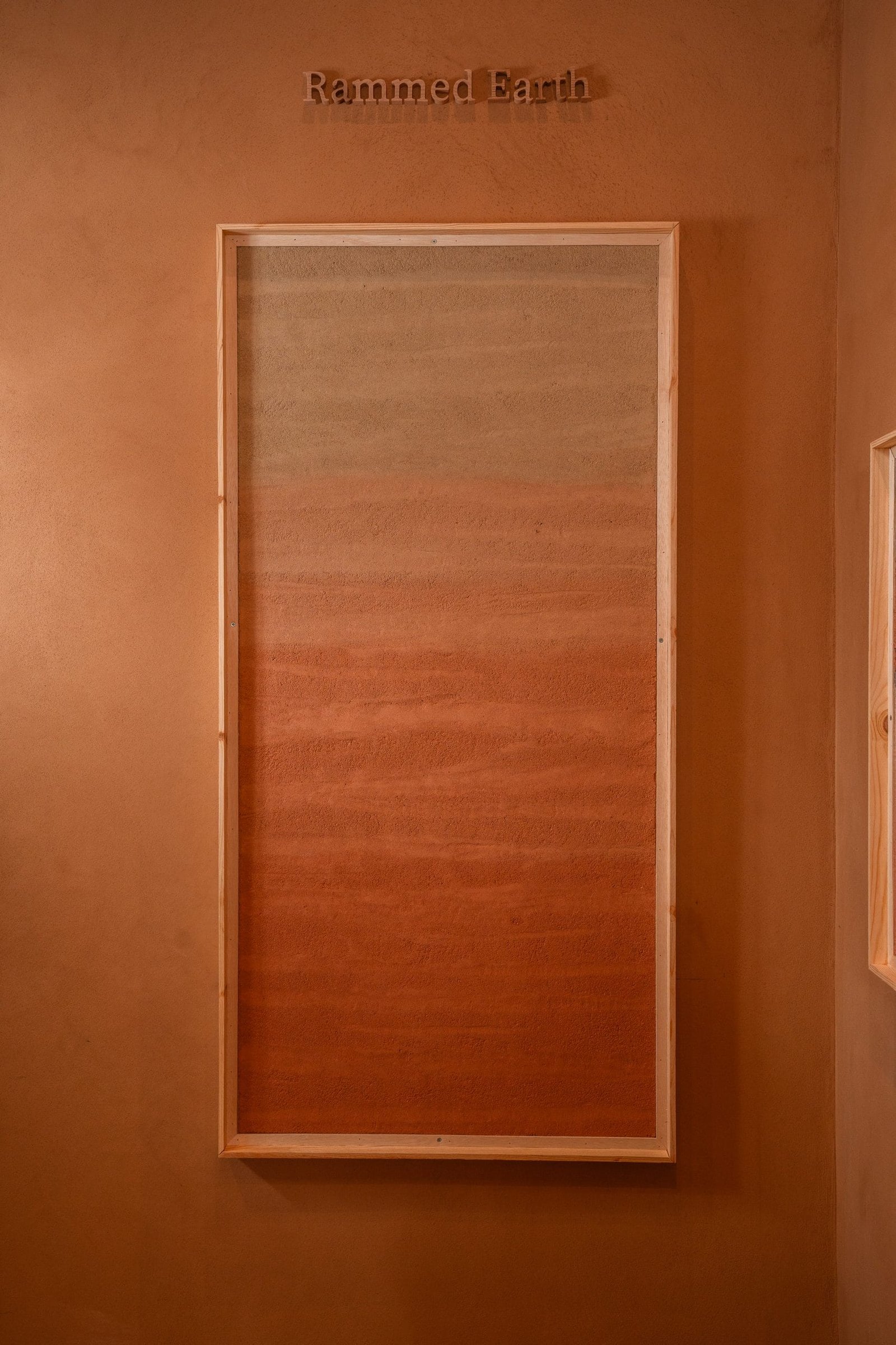 Framed rammed earth plaster panel under the Rammed Earth label at the Conmarble showroom, graded grey through ochre to terracotta.