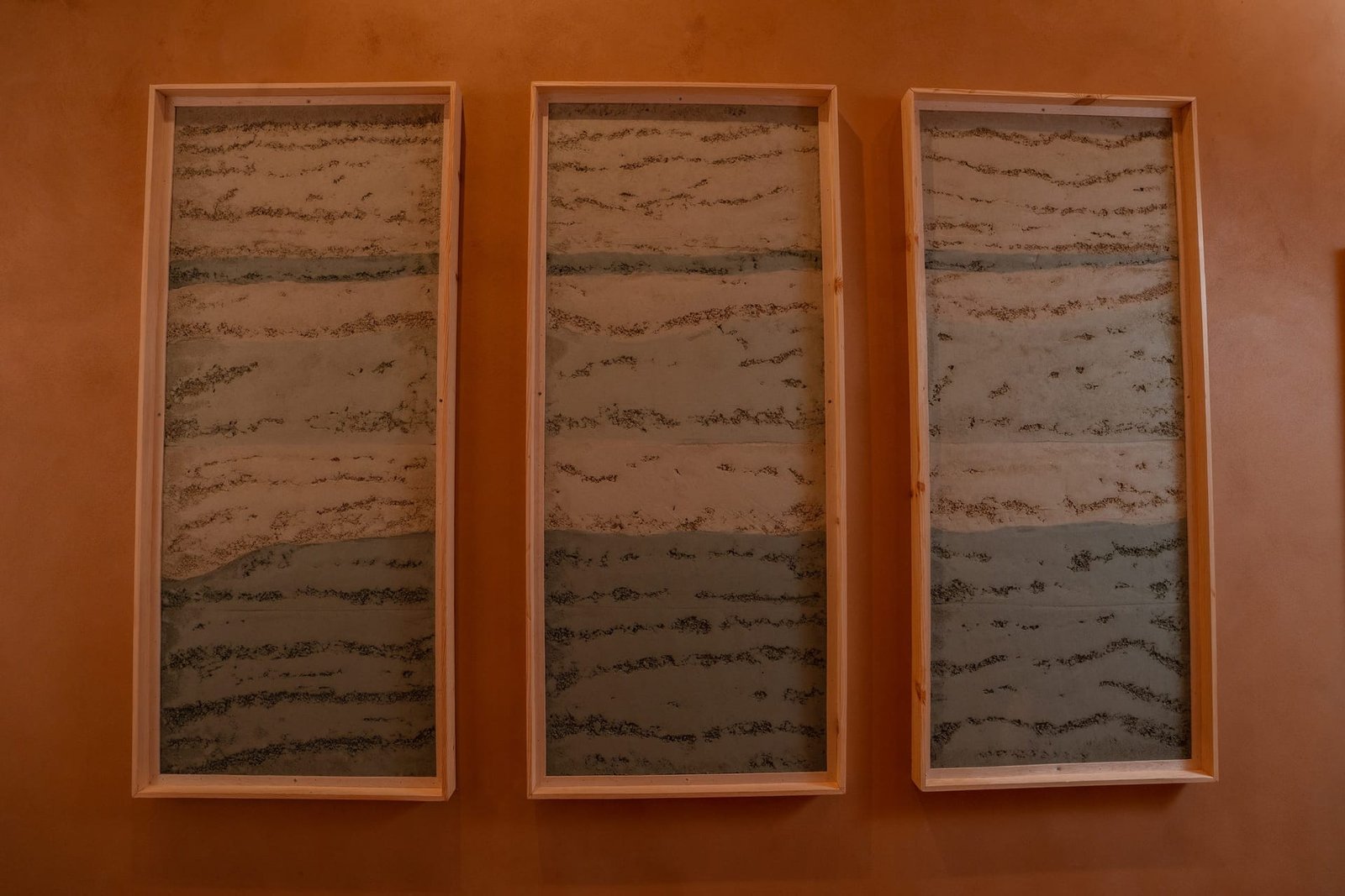 Three tall framed rammed earth plaster panels at the Conmarble showroom, cream and grey-green bands with pronounced open grain.